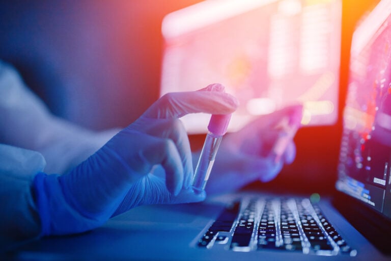 Gloved hands holding vials near a keyboard and computer monitor in a dark setting
